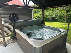 Un jacuzzi con muelle de madera y techo metálico, rodeado de césped y árboles.