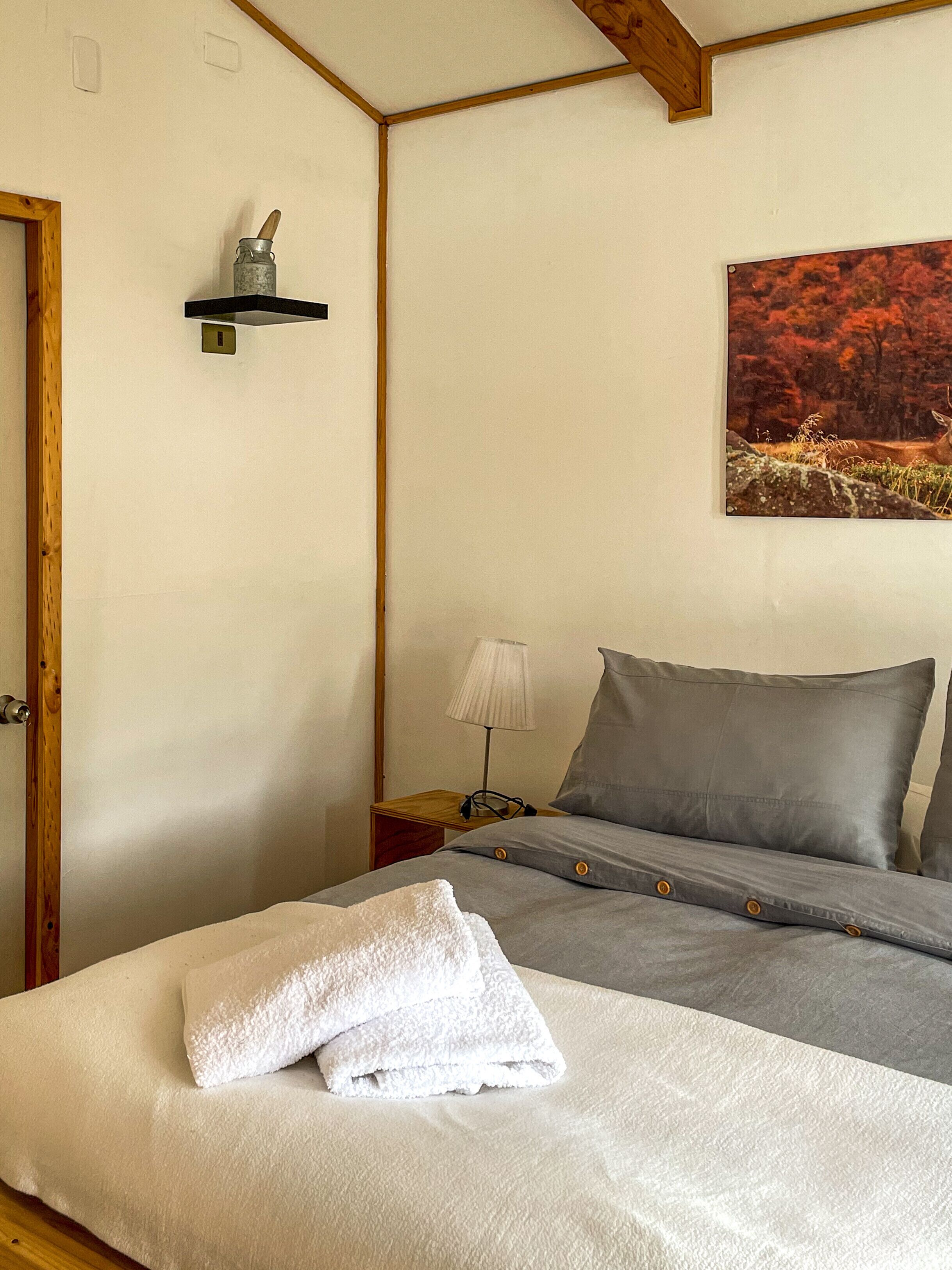 A bedroom with a bed, a nightstand, a lamp, a painting, and a wooden ceiling beam.