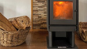 Una estufa de leña con un fondo de pared de piedra, una cesta de mimbre con leña y una silla de mimbre.