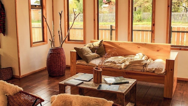 Una habitación acogedora con un sofá de madera, una mesita de centro con libros y ventanas amplias con vistas a una valla de madera y follaje.