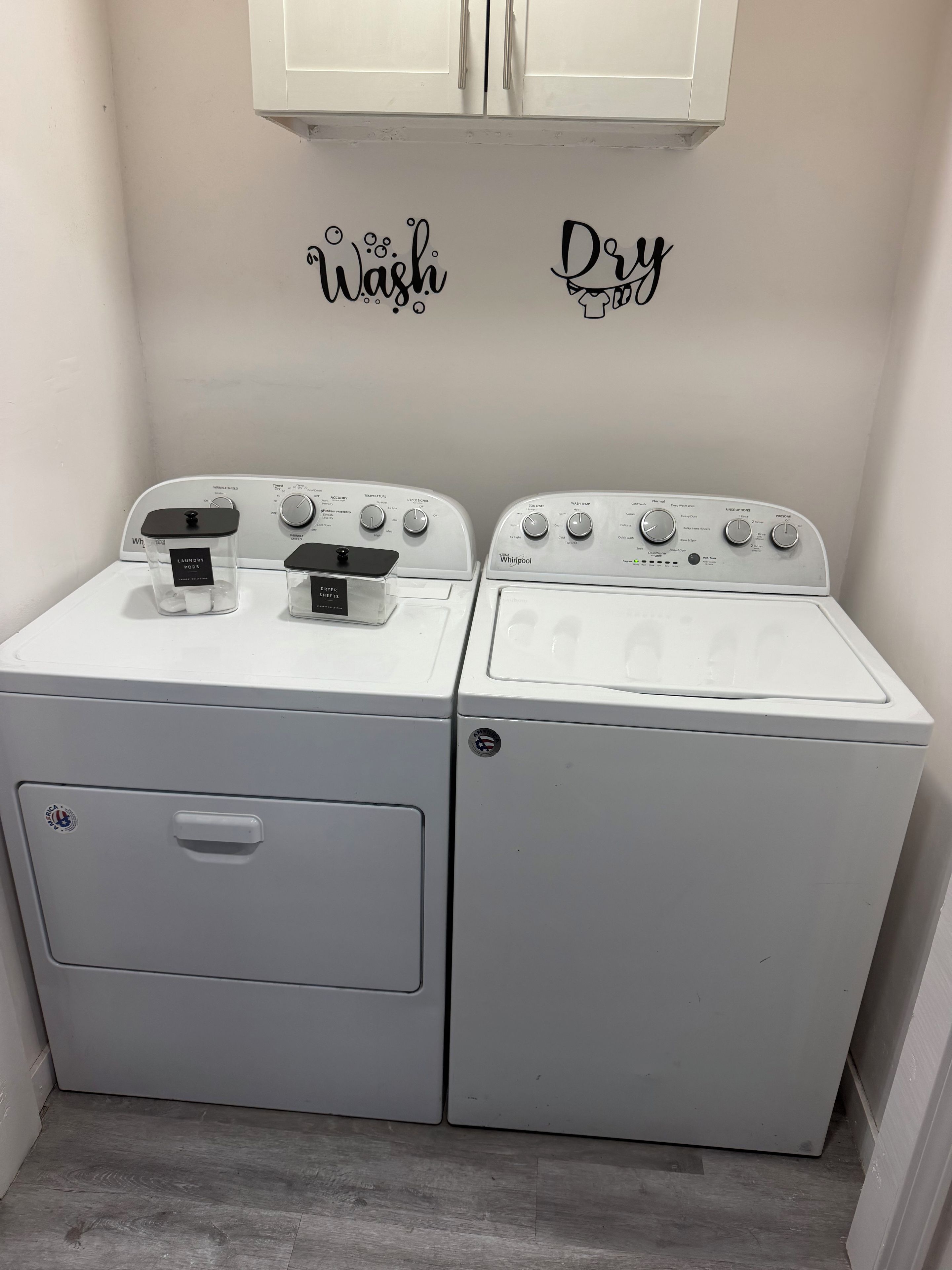 A laundry room with two white washing machines, a cabinet above, and wall decals saying 'Wash' and 'Dry'.