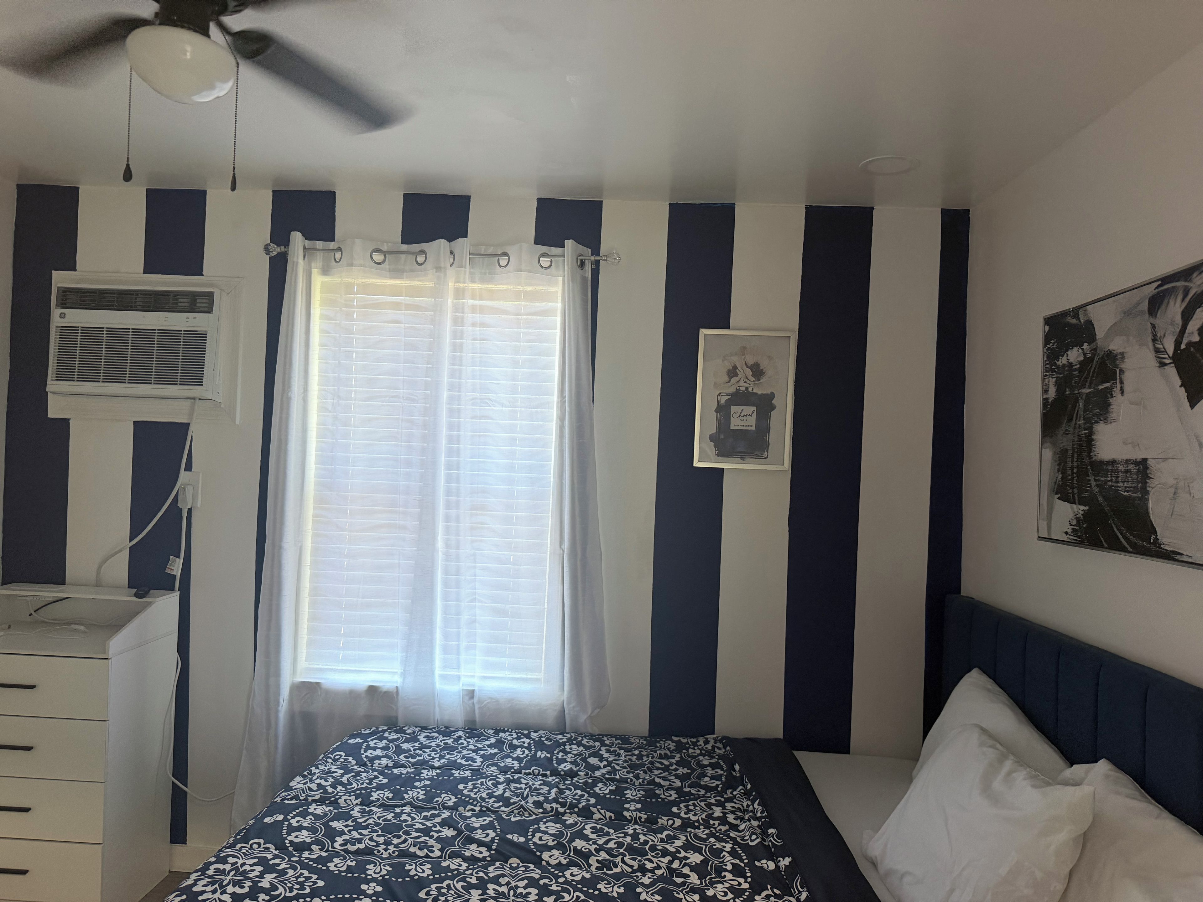 Een slaapkamer met een bed, een plafondventilator, een raam met gordijnen, een wandairconditioner en een ingelijst beeld.
