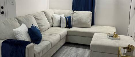 Ein Wohnzimmer mit Ecksofa, einem kleinen Hocker, einem Flachbildfernseher und Kunstwerken an den Wänden.