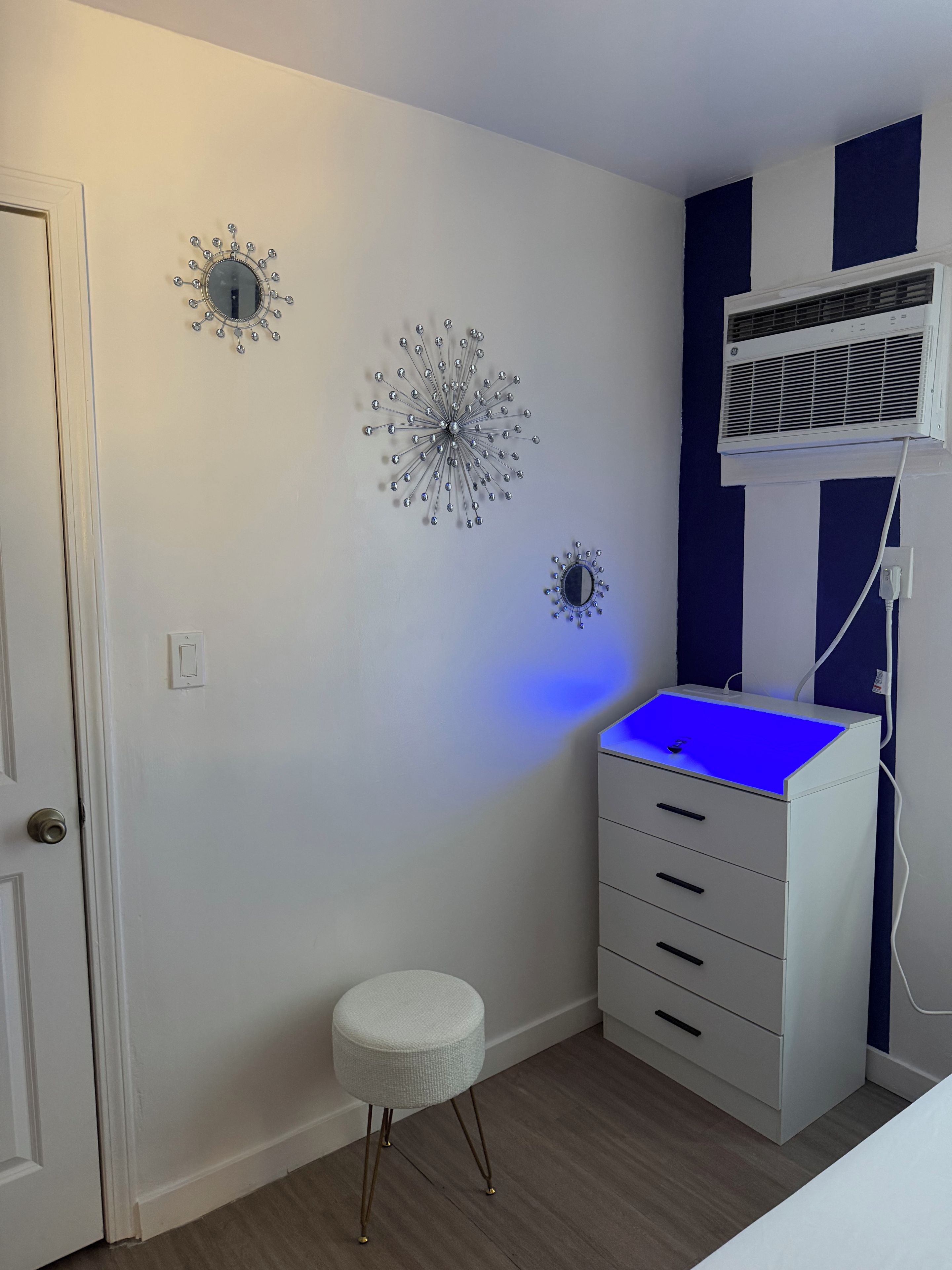 A room with a white wall, a blue accent wall, a white cabinet with drawers, a small stool, and a wall-mounted air conditioner.
