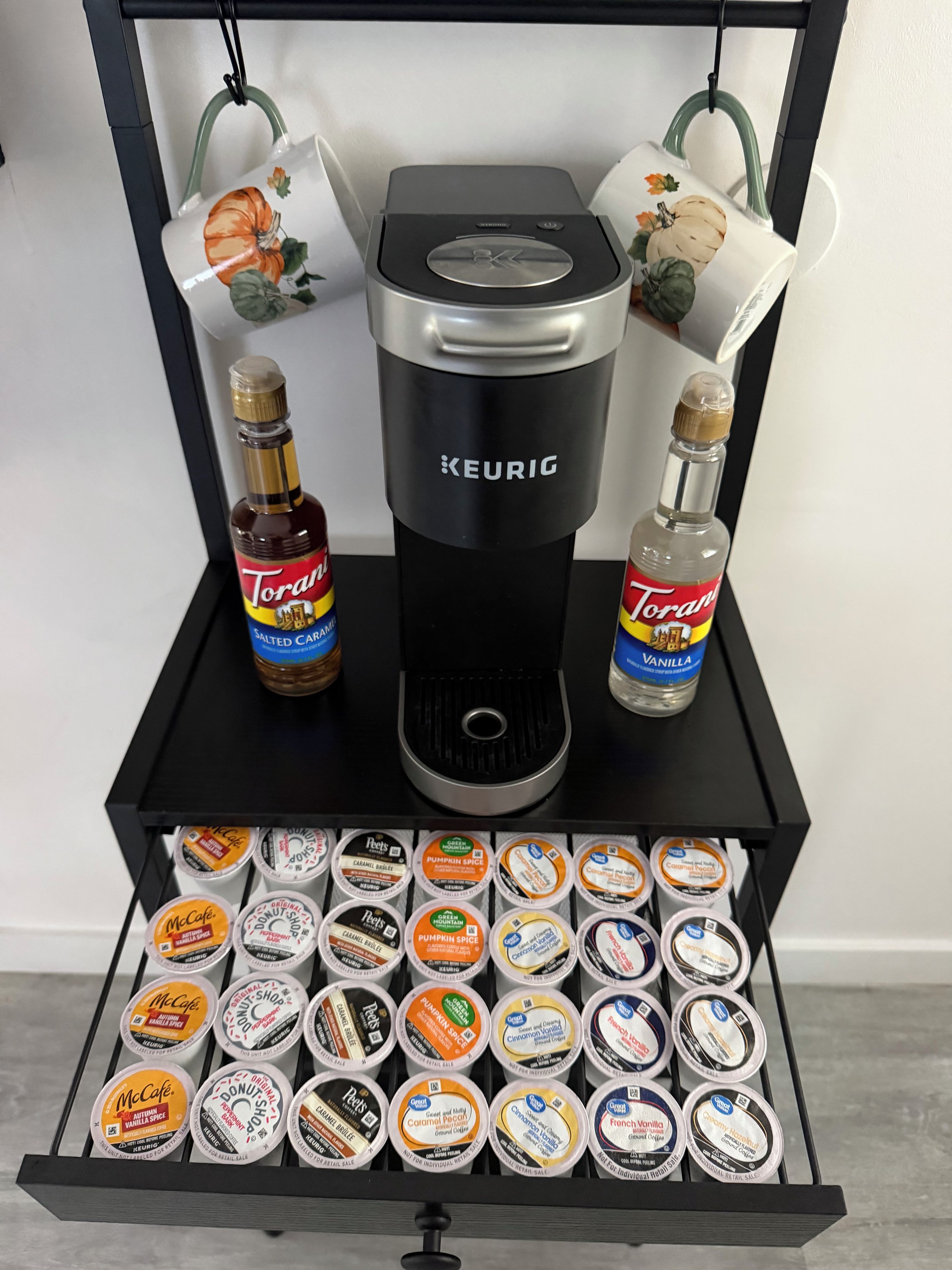 Eine Kaffeemaschine mit schwarzem Sockel und silberner Oberseite, zwei Flaschen Torani-Sirup und eine Schublade mit verschiedenen McCafé-Aromen.