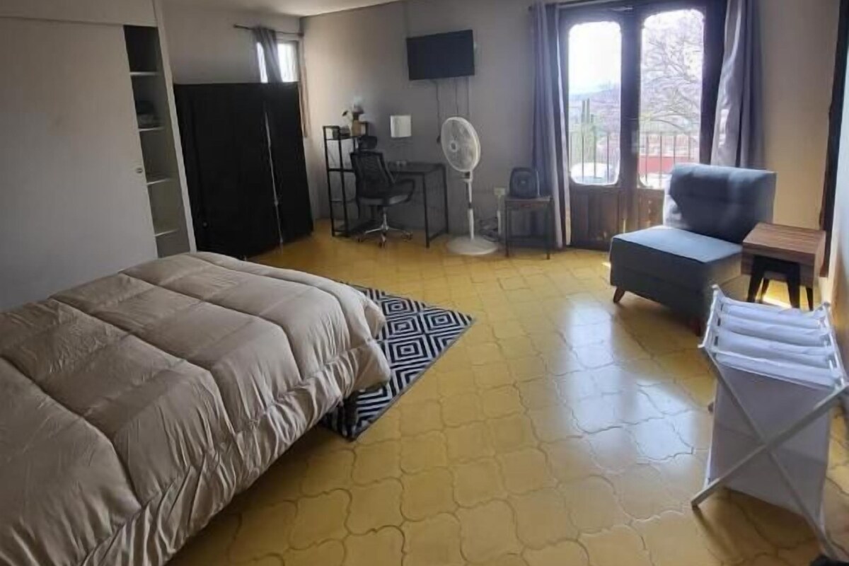 Une chambre d’hôtel avec un lit, un bureau, une chaise, un ventilateur et une fenêtre avec des rideaux.