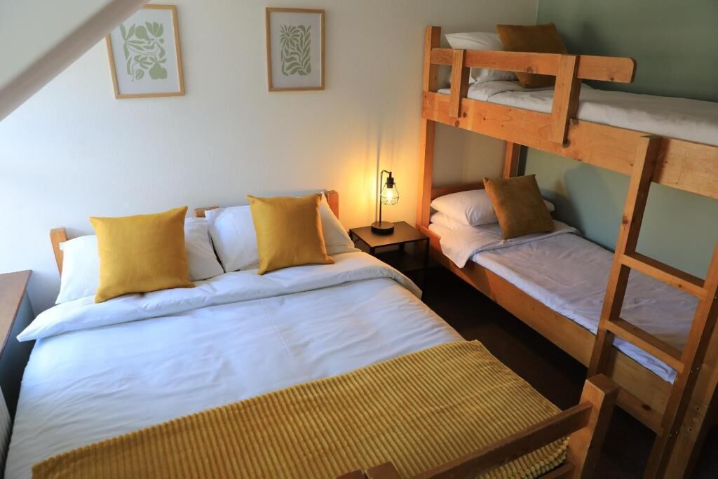Ein Schlafraum mit Etagenbett, einem Einzelbett, zwei Kissen, einer Nachttischlampe und zwei gerahmten Bildern an der Wand.