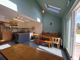 Una habitación con una mesa de madera, bancos, una pequeña isla con un lavabo y un ventanal grande.
