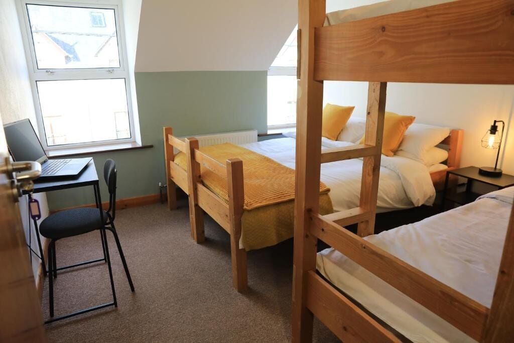 Habitación tipo dormitorio con literas, un escritorio con una laptop, una silla y una mesita de noche con una lámpara.