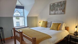 A bedroom with a sloped ceiling, a bed with white bedding and yellow pillows, a wooden headboard, a bedside table with a lamp, and a painting on the wall.
