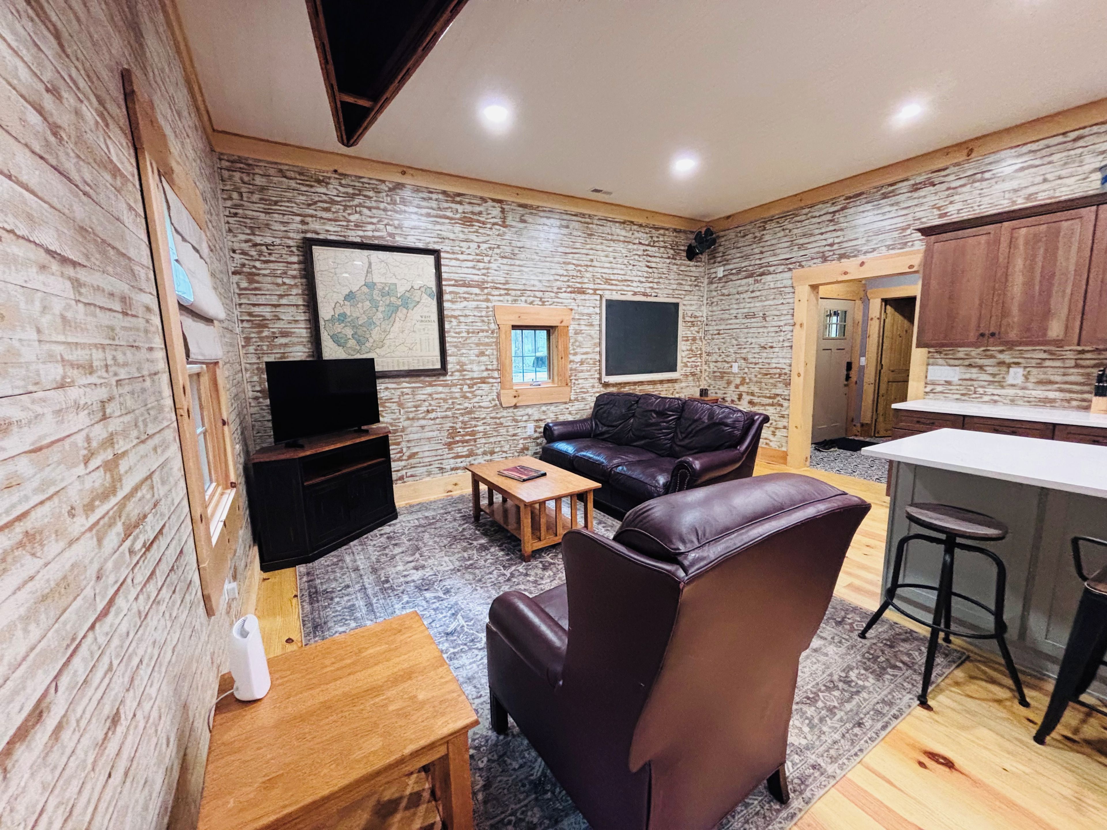 A cozy living room with a leather sofa, a coffee table, and a TV. There is a kitchen area with wooden cabinets and a bar stool.