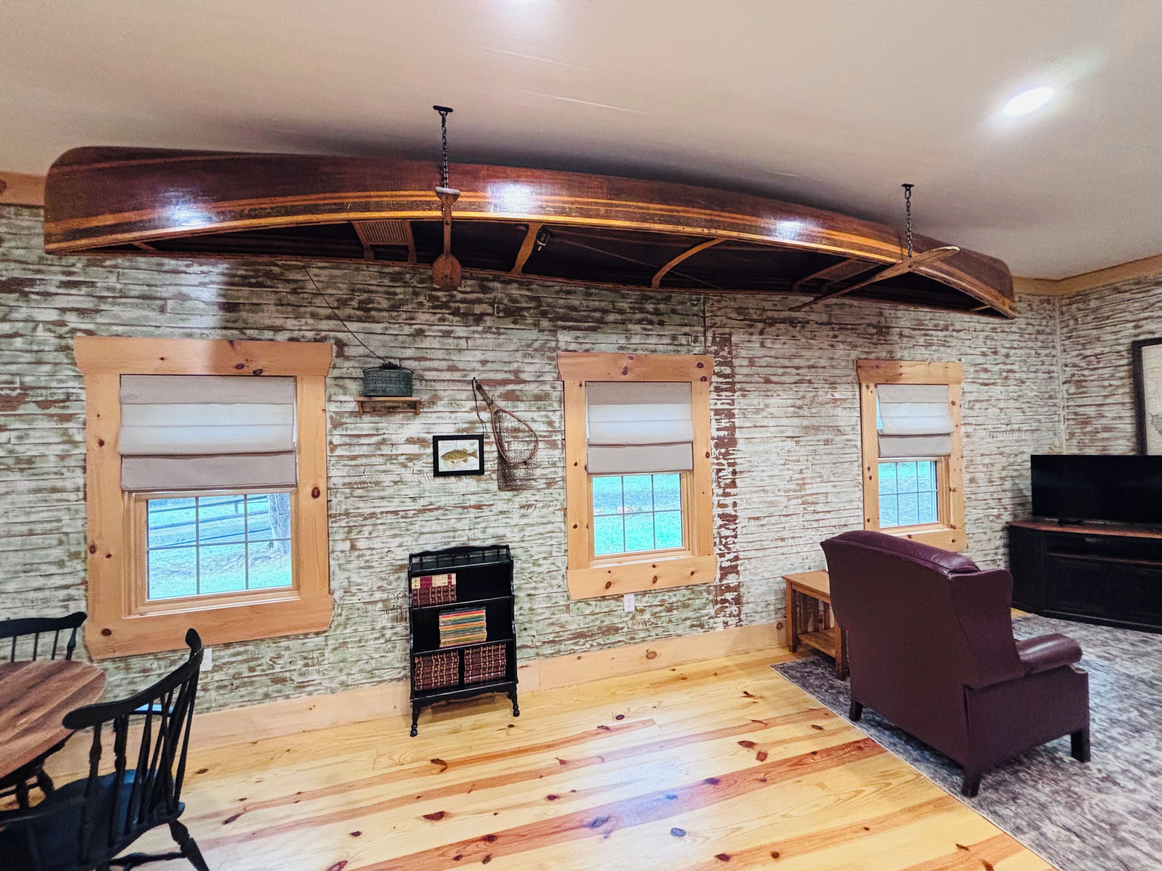 A room with a rustic wooden ceiling, two windows with blinds, a television, a sofa, and a small bookshelf.