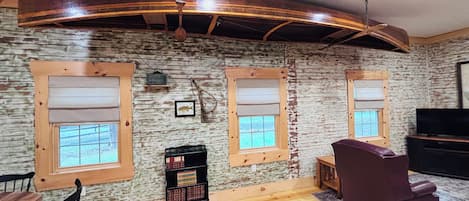 A room with a rustic wooden ceiling, two windows with blinds, a television, a sofa, and a small bookshelf.