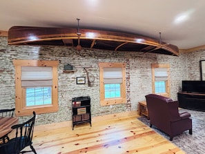 A room with a rustic wooden ceiling, two windows with blinds, a television, a sofa, and a small bookshelf. - Historic Schoolhouse/Log Cabin - minutes to the beautiful South Branch River! (Romney)