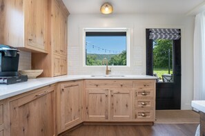 Ett kök med träskåp, en vit bänkskiva, ett fönster med utsikt över grönområden och en svart dörr med en mönstrad gardin.