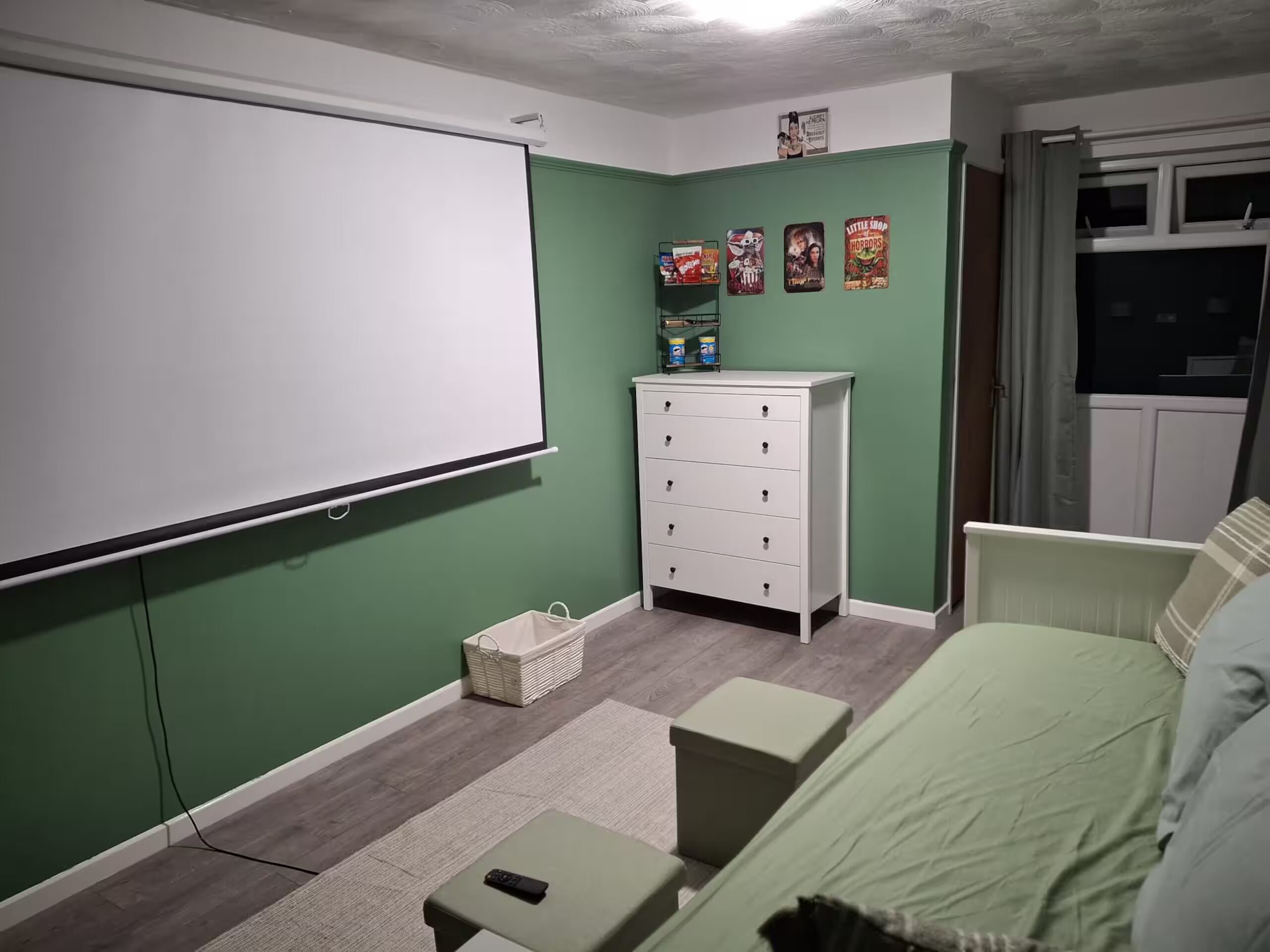 A room with a projector screen, a white dresser, a green sofa, and a small table with a remote control.