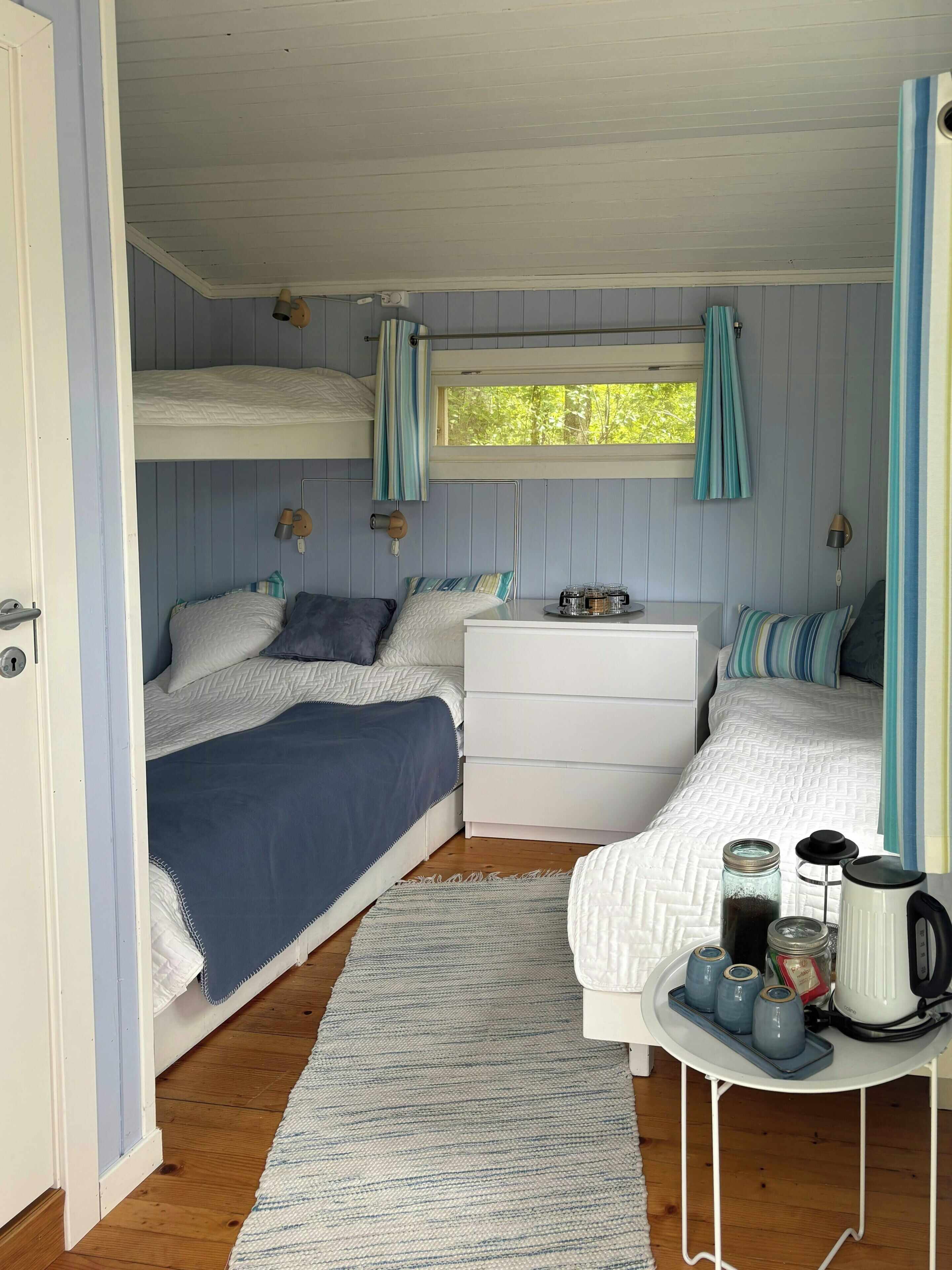 A compact bedroom with bunk beds, a bedside table with a kettle and cups, and a window with curtains.