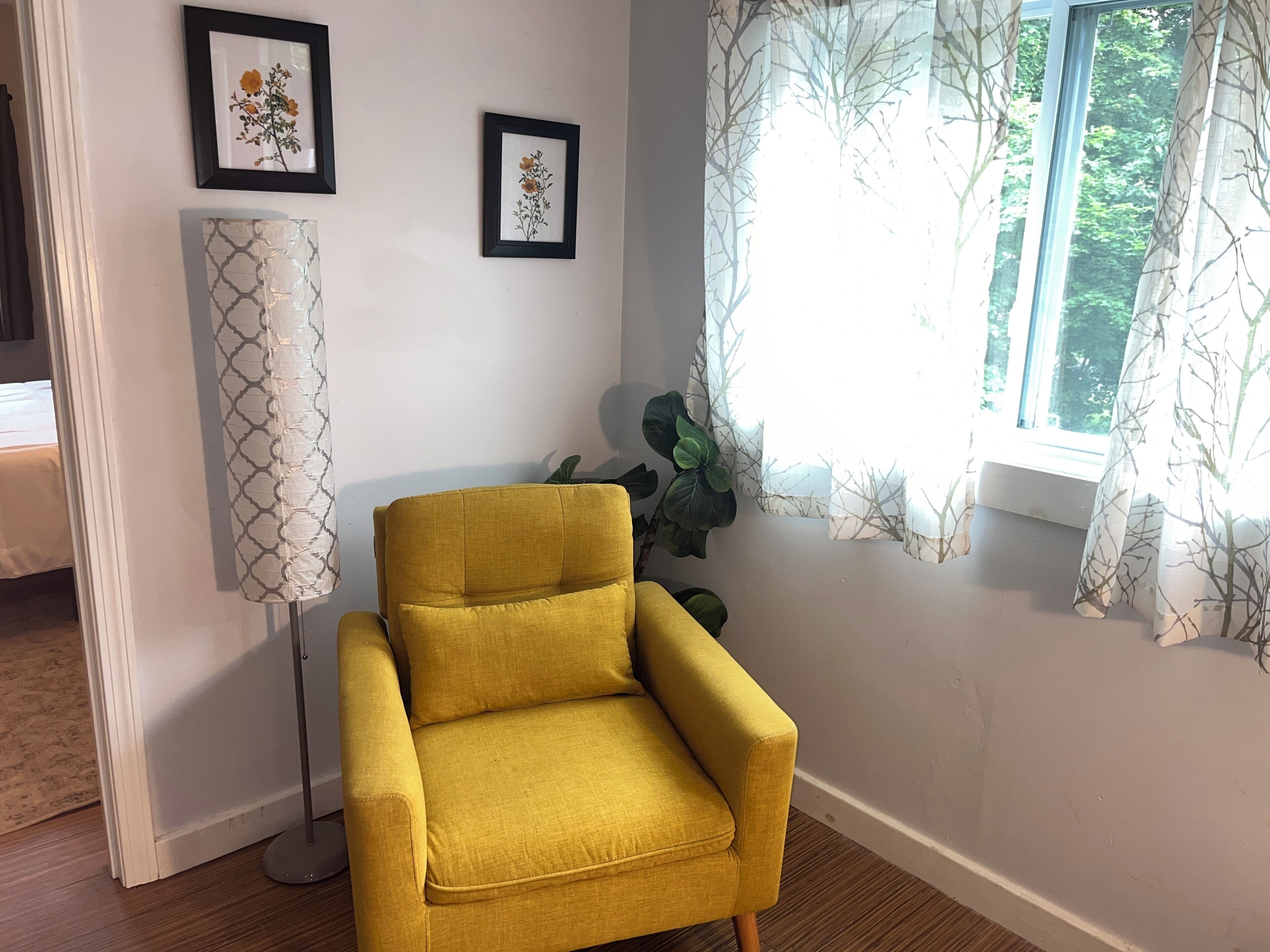 Un fauteuil jaune dans une pièce avec une fenêtre, un lampadaire et deux tableaux encadrés au mur.
