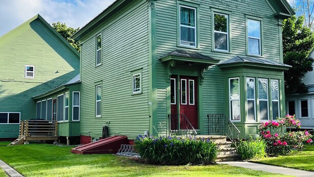 A two-story house with a red door, a front porch, and a small front yard with a wooden deck and flowers.