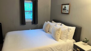 A neatly made bed with white bedding and several pillows, a bedside table with a plant, and a framed picture on the wall.