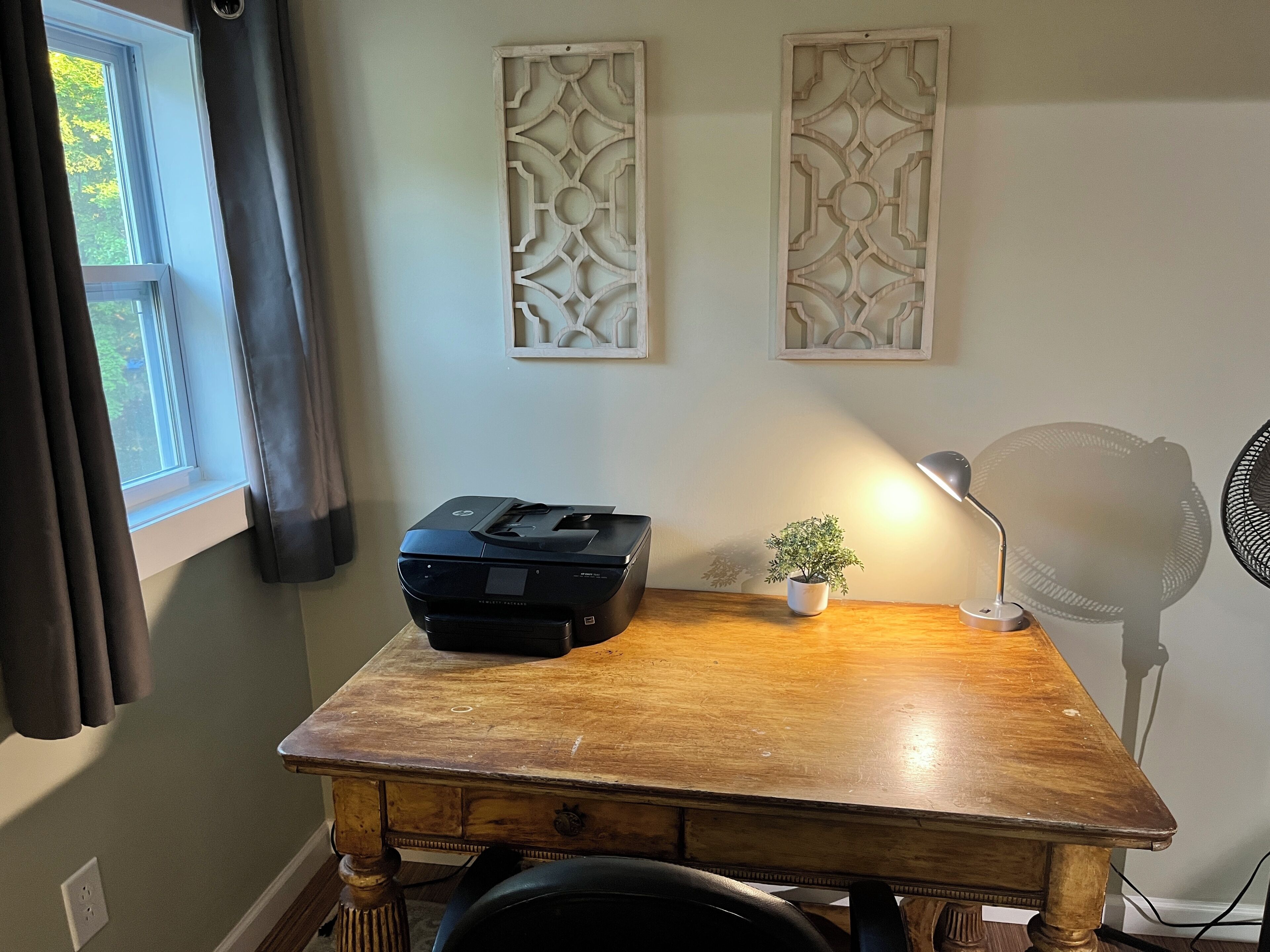 Un bureau en bois avec une imprimante, une petite plante en pot, une lampe de bureau et un ventilateur sur pied.