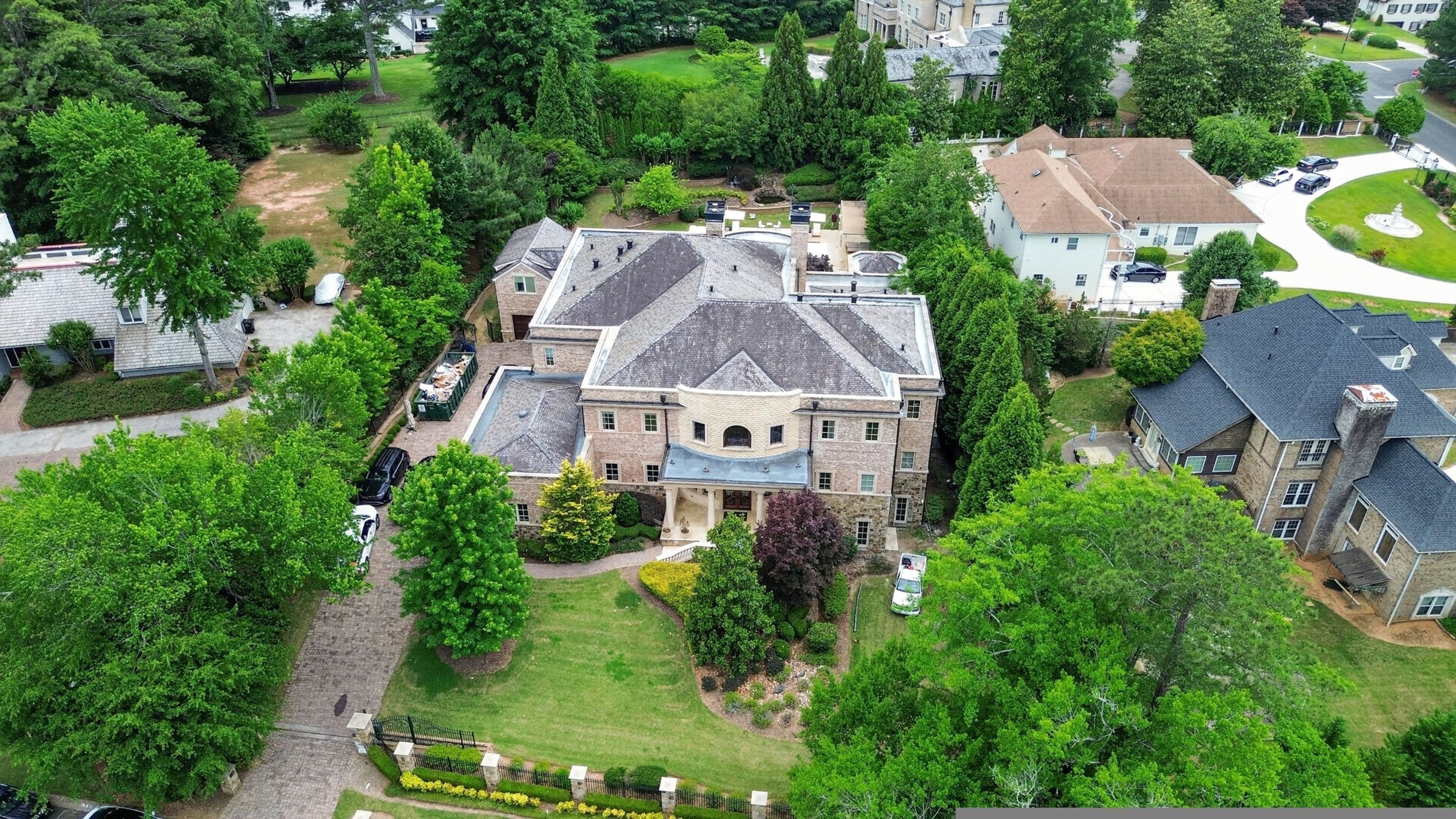 Een groot, meerlaags huis met een opvallend centraal gedeelte, omringd door weelderige begroeiing en andere woongebouwen.