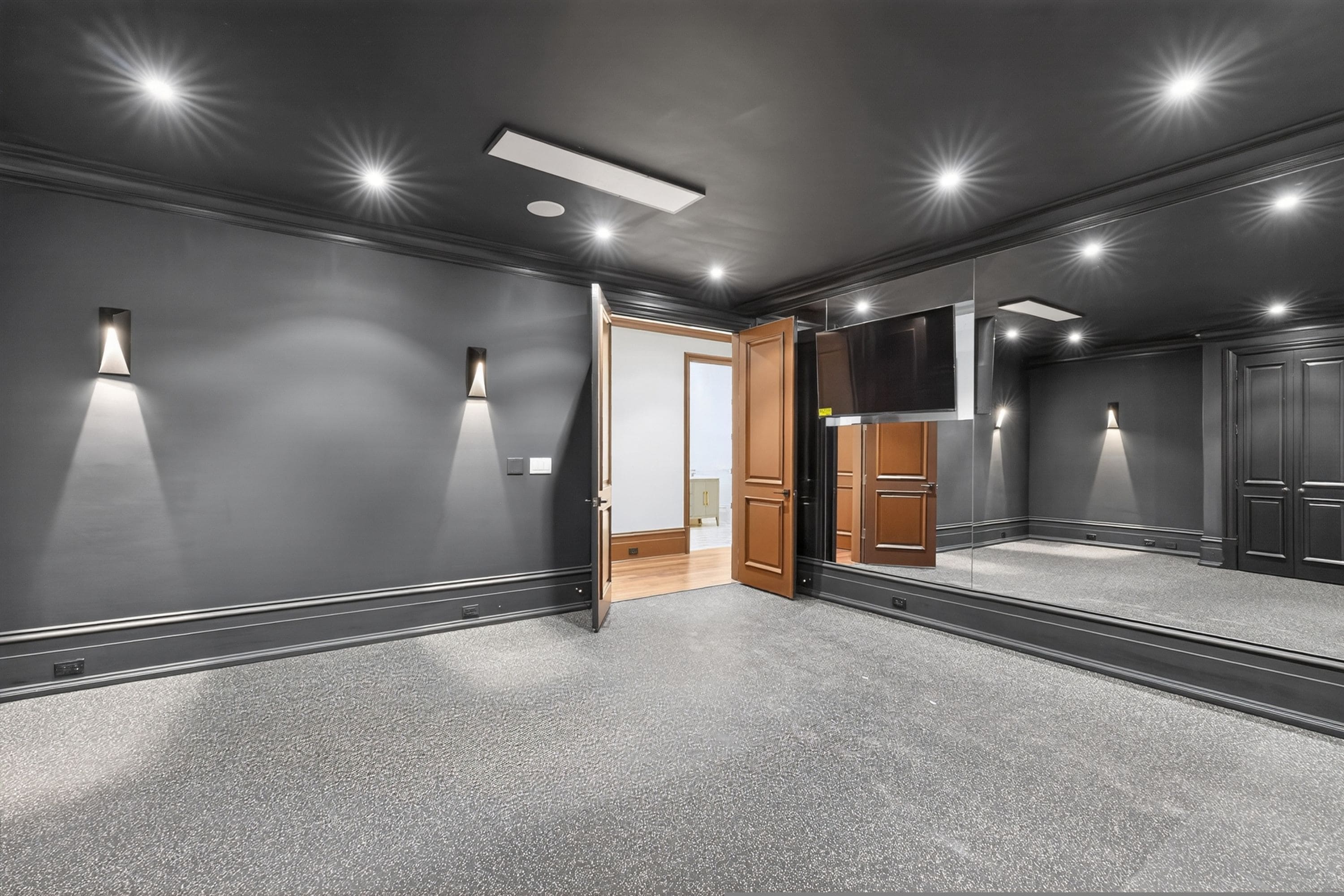 Una habitación con un espejo grande, luces empotradas en el techo y una puerta que da a otra estancia.