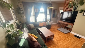 Een woonkamer met een groene bank, een houten salontafel, een flatscreen televisie en een groot raam met gordijnen.