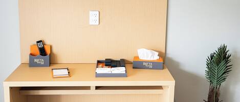 A hotel room with a wooden desk, a flat-screen TV mounted above it, a power outlet, and a small potted plant.