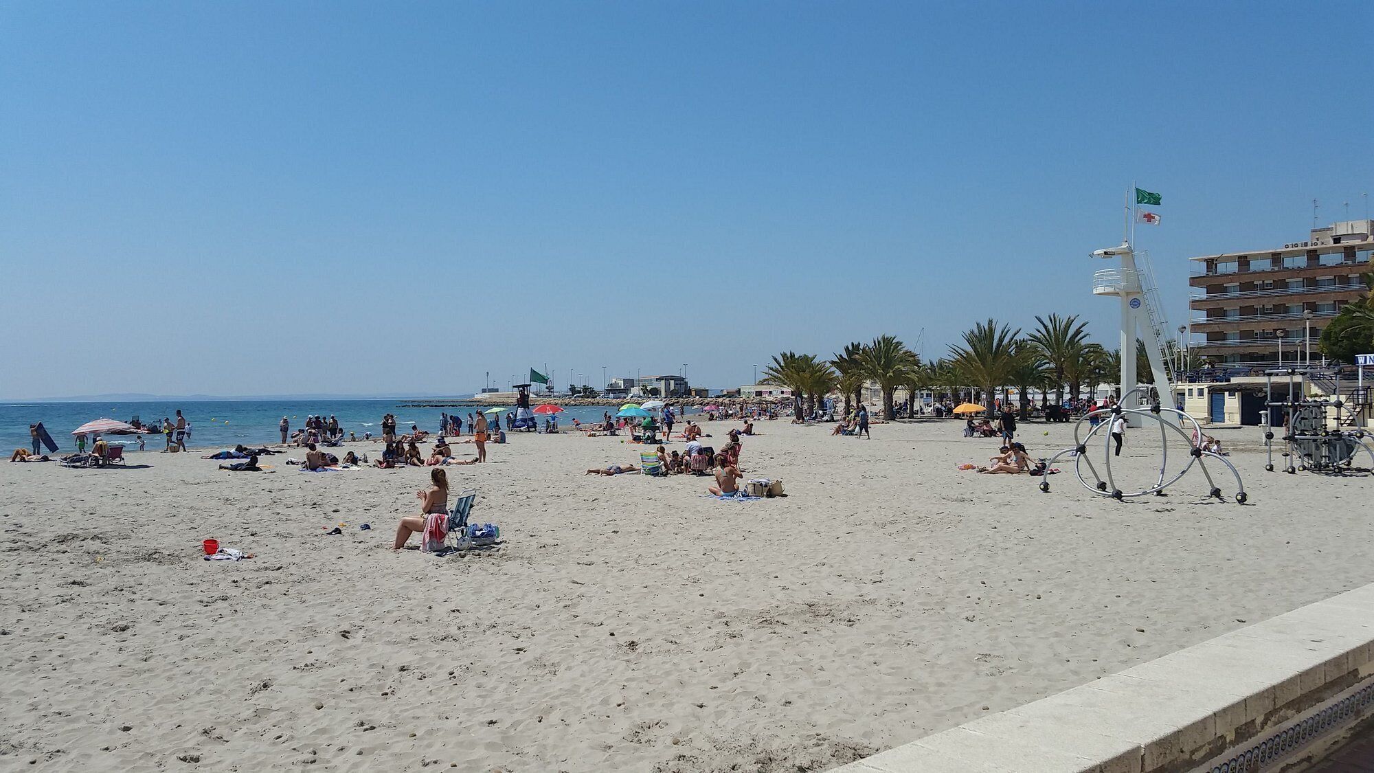 A beach with people sunbathing and walking, a lifeguard tower, and a building with balconies.