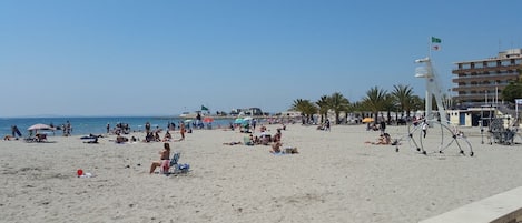 A beach with people sunbathing and walking, a lifeguard tower, and a building with balconies.