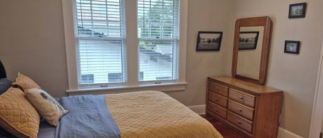 A bedroom with a bed, a dresser, a window with blinds, and a mirror on the wall.