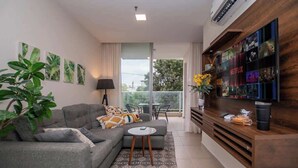 Ein modernes Wohnzimmer mit grauem Sofa, einem Couchtisch, einem an einer Holzwand montierten Fernseher und einem großen Fenster mit Blick ins Grüne.