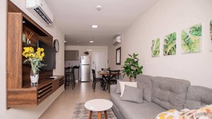 Ein modernes Wohnzimmer mit einem grauen Sofa, einem weißen Couchtisch und einem Essbereich, im Hintergrund ist eine Küche zu sehen.