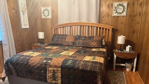 Ein Bett mit Decke, einem Holzkopfstück, zwei Nachttischen und einem Beistelltischchen mit Lampe.