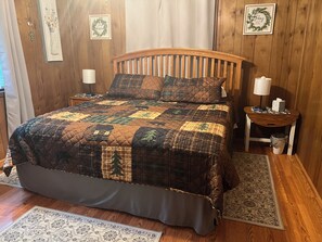 Ein Bett mit Decke, einem Holzkopfstück, zwei Nachttischen und einem Beistelltischchen mit Lampe.