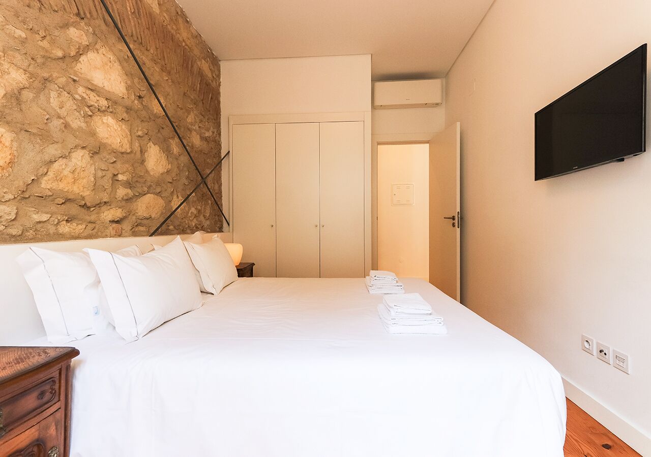 A hotel room with a bed, a wooden nightstand, a stone wall, a wardrobe, and a flat-screen TV.