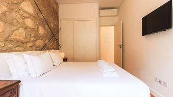 A hotel room with a bed, a wooden nightstand, a stone wall, a wardrobe, and a flat-screen TV.