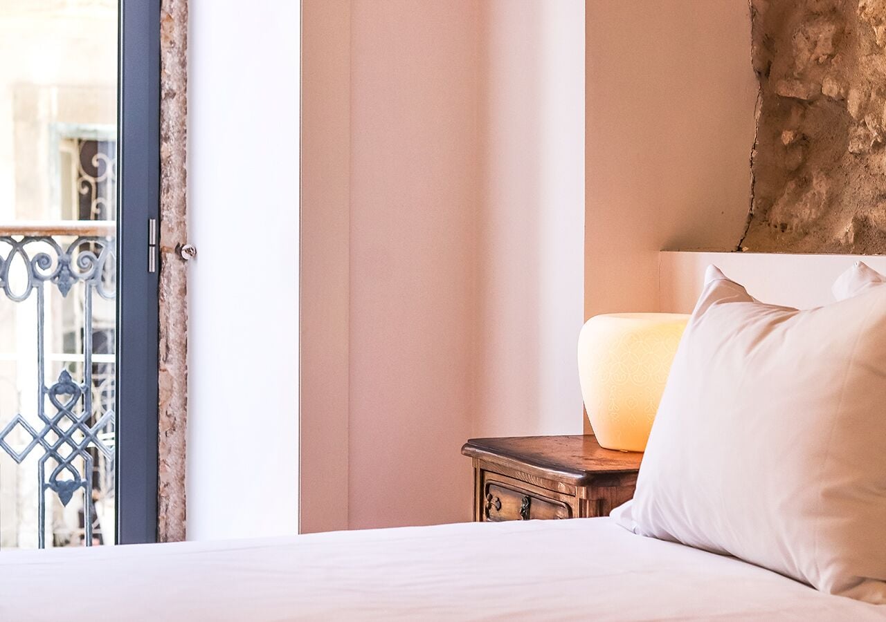 A hotel room with a bed, a nightstand with a lamp, and a view of a balcony with a metal railing.