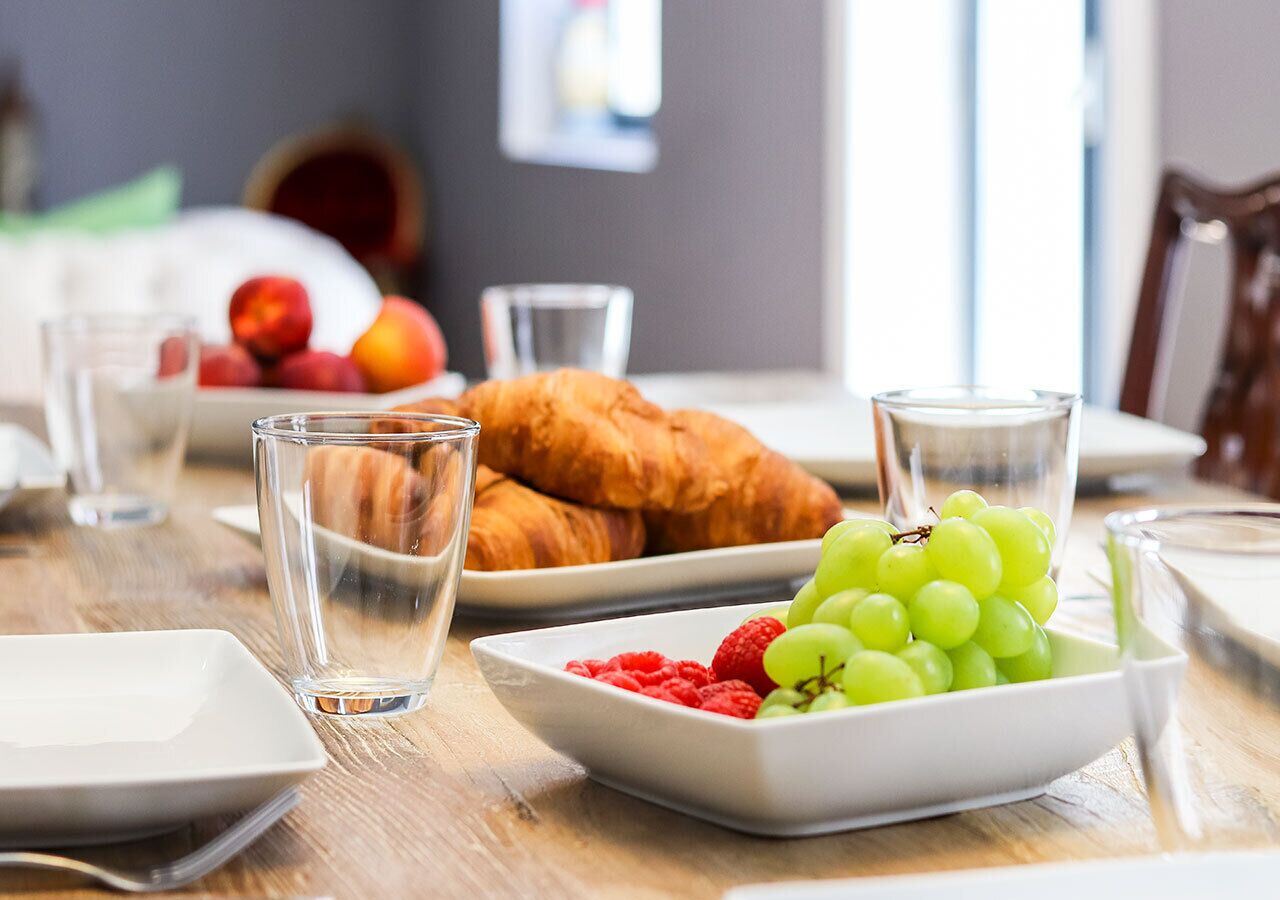 A breakfast setting with croissants, grapes, raspberries, and water glasses on a wooden table.