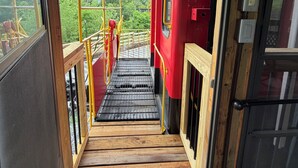 A wooden staircase leading to a red train carriage with a view of greenery outside.
