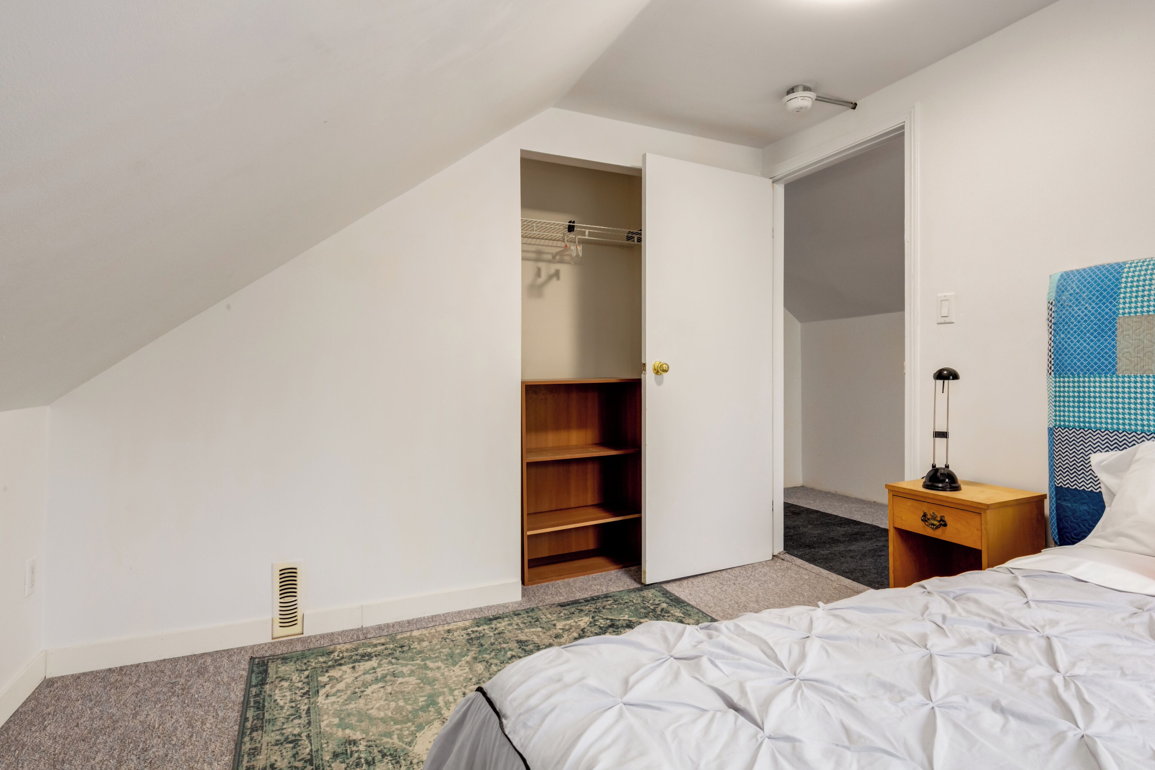 A bedroom with a bed, a nightstand, a lamp, a wardrobe, and a door leading to another room.