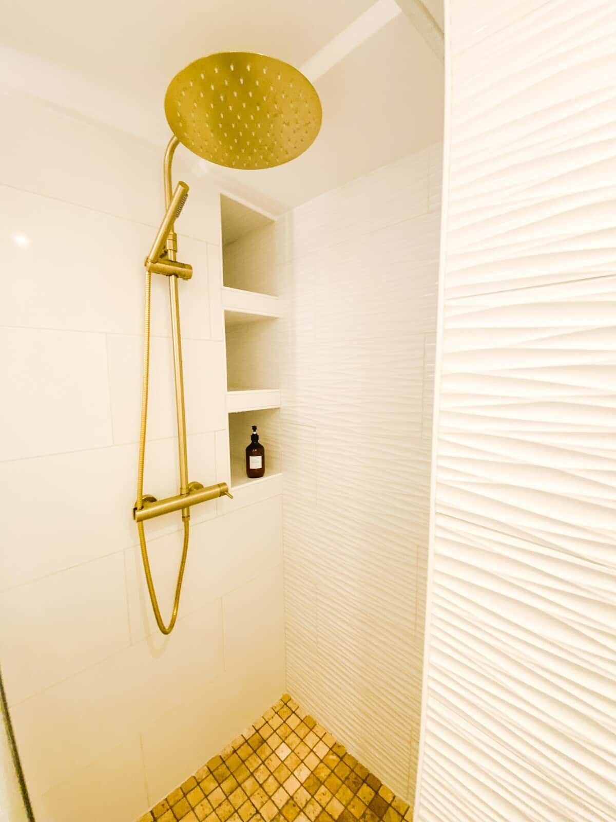 A shower with a gold showerhead and a bottle of shampoo on a shelf.