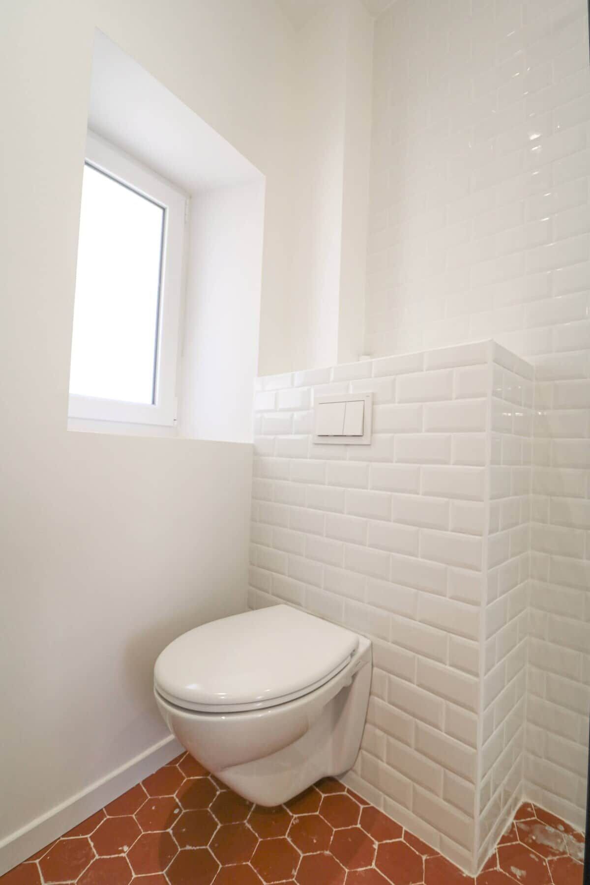 Une salle de bain avec des toilettes blanches, des murs recouverts de carreaux blancs, une fenêtre et un sol carrelé avec des motifs hexagonaux.