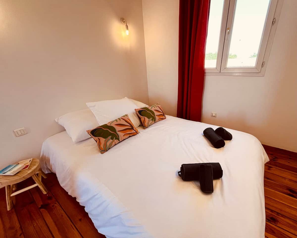 Una cama bien hecha con ropa de cama blanca y almohadas decorativas, una mesita de noche de madera con una revista y una ventana con cortinas rojas.