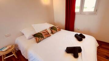 Una cama bien hecha con ropa de cama blanca y almohadas decorativas, una mesita de noche de madera con una revista y una ventana con cortinas rojas.