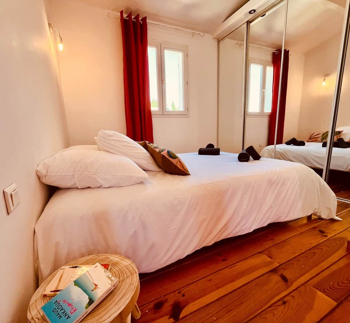 A bedroom with a bed, a bedside table with books, a mirror, and a window with curtains.