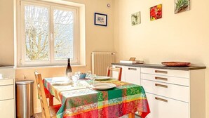 Ein Essbereich mit einem Tisch für vier Personen, ein Fenster mit Jalousien, ein Heizkörper und ein Küchenbereich mit Schränken und einer Arbeitsfläche.