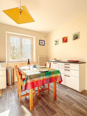 Ein Essbereich mit einem Tisch für vier Personen, ein Fenster mit Jalousien, ein Heizkörper und ein Küchenbereich mit Schränken und einer Arbeitsfläche.