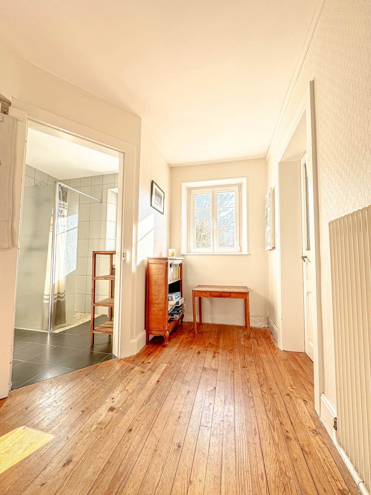 A hallway with wooden flooring, a radiator, a window, and a door leading to a bathroom.
